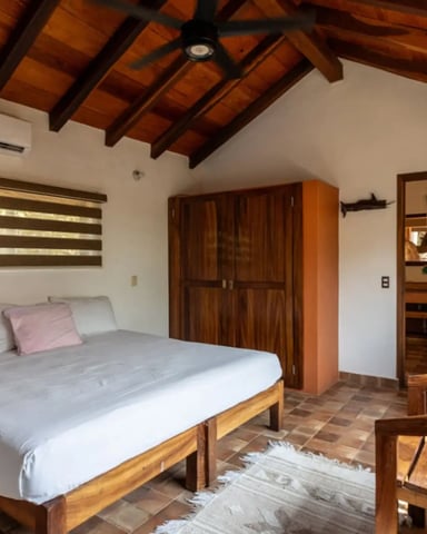 Bedroom with wooden bed frame, exposed beam ceiling, wooden wardrobe, and ensuite bathroom visible through open door