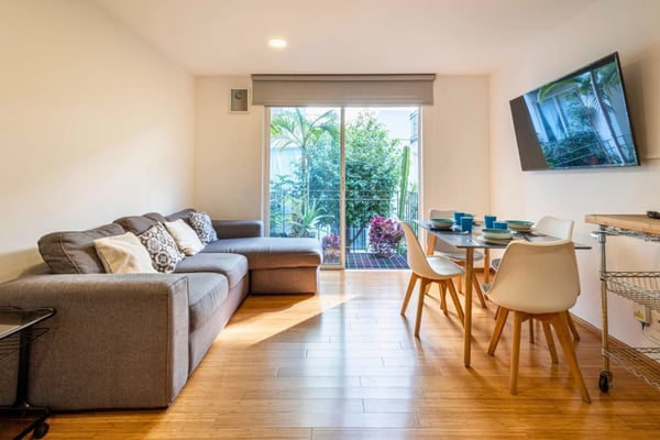 Open-plan living room with gray sofa, dining table, wall-mounted TV, and glass doors opening to balcony with tropical plants