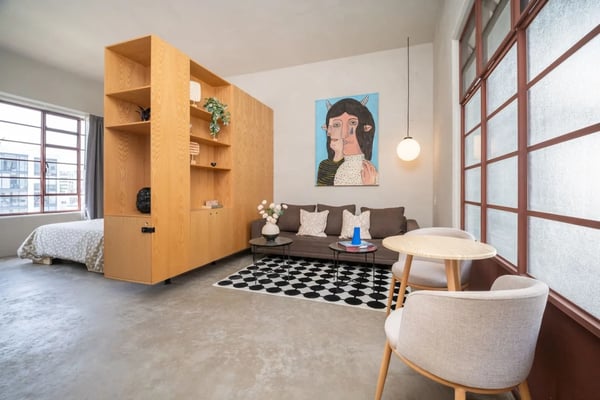 Studio apartment with wooden room divider, brown sofa, two cream armchairs, round table, and city views from large windows