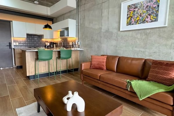 Cocina y sala de estar de planta abierta con gabinetes de madera, asientos en la isla y sofá gris con cojines decorativos amarillos