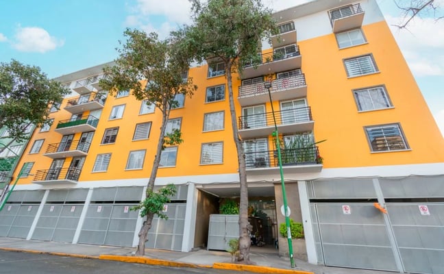 Modern apartment building with bright yellow and white facade, multiple balconies, ground-floor garage, and street trees.